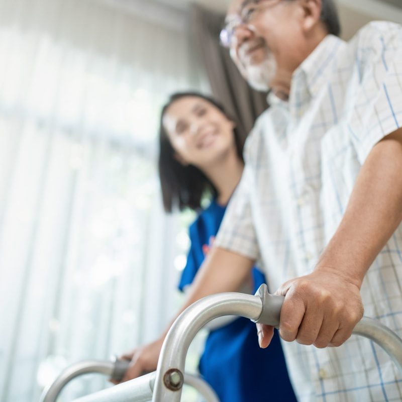 Close up of nurse help disable man walk with walker at nursing home. Caregiver therapist doctor support senior older handicapped patient use Zimmer frame in house. Medical therapy insurance concept.